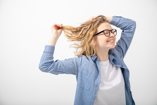 Side view of young smiling beauteous woman with closed eyes raising hands, holding short fair wavy hair, touching head, wearing glasses, denim shirt, white T-shirt on white background. Copy space.