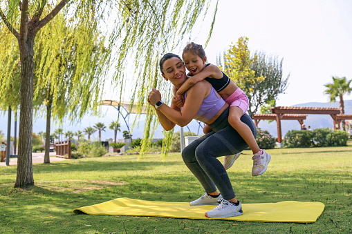 Mother and daughter spending quality time together, bonding through physical exercise. Little girl joined her mom for a morning exercise, doing squats in the park. Close up, copy space, background.