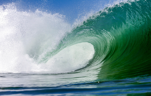 Closeup horizontal photo of a curving blue wave with the wind blowing spray from the top, lit up by the morning sun