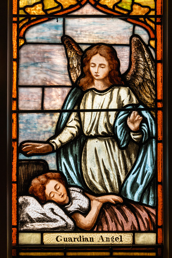 A stained glass window from a Catholic Church that is of an angel guarding a small child sleeping in her bed.
