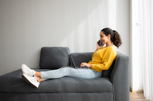 Side view of a happy young woman using laptop and smartphone while sitting on sofa at home