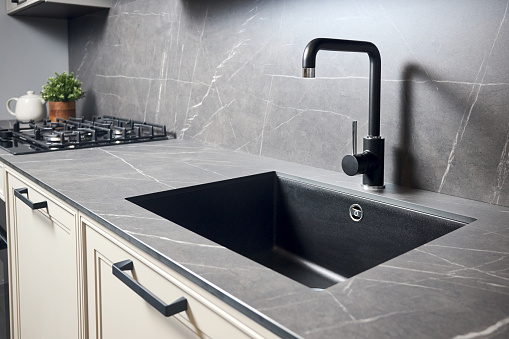 Compact undermount sink. Kitchen sink area with black square matte sink tap in contemporary style. Matte black and stoneware kitchen design. Black ceramic sink with gas hob and oven, in background.