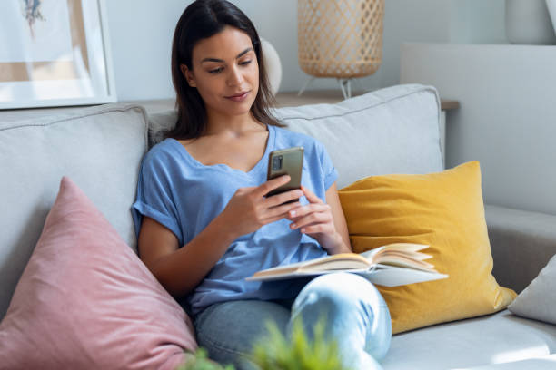 istock Pretty young woman using her mobile phone while reading a book on sofa at home. 1690935120