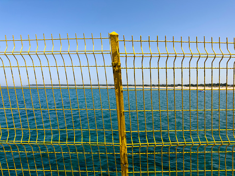 Blue sky behind a wire fence. Beach and sea in the background. Sunny summer day. Yellow wire mesh for security on the ferry pier. No people.