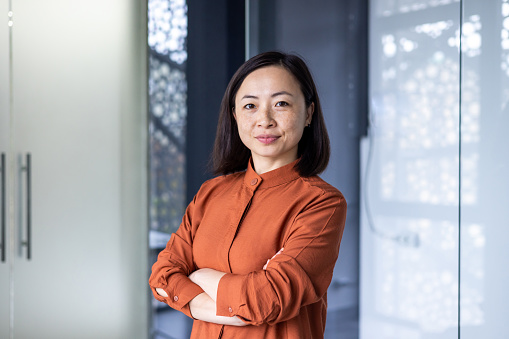 Portrait of a young beautiful confident Asian businesswoman, standing in an office center with her arms crossed, on her chest and looking seriously at the camera.
