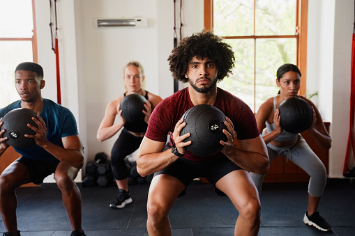 Four focused multiracial young adults doing medicine ball squats at the gym