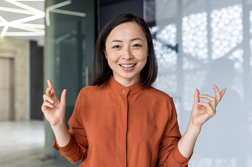Young beautiful, successful Asian businesswoman, portrait of female worker inside office smiling and looking at camera, online video call, remote meeting with colleagues and partners.