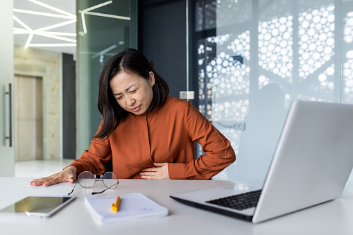 Sick woman at workplace has acute stomach pain, asian business woman holding hand on stomach from pain, asian woman working sitting at desk with laptop inside office.