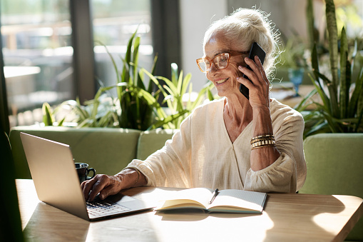 Smiling senior woman talking on mobile phone and typing on laptop while sitting at table in cafe