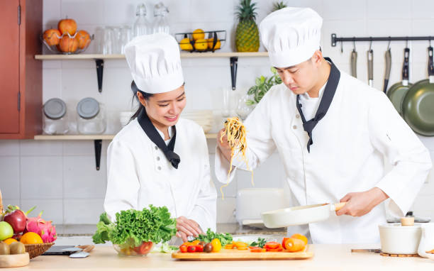 istock Two Asian professional couple chef wearing white uniform, hat, helping each other, preparing meal,cooking in kitchen together, smiling with happiness and confidence. Food Concept. 1690942616