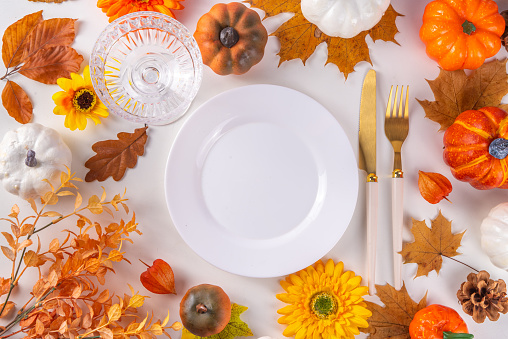 Autumn holiday table setting. Thanksgiving holiday menu, greeting card. White kitchen table with plate and cutlery, fall leaves, flowers and decor flat lay top view copy space