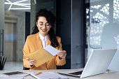 Happy young Latin American woman received bill with earned salary, cash reward, good news. He is sitting at the desk in the office, holding an envelope with a letter in his hands.