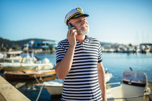 Mature man standing near the sea dressed in a sailor's shirt and hat using smart phone