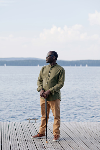 Vertical image of senior man with stick standing on pier against the lake