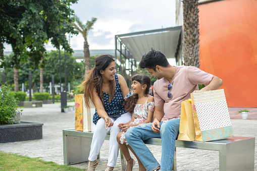 Happy young parents with daughter talking while sitting on bench outside shopping mall in city