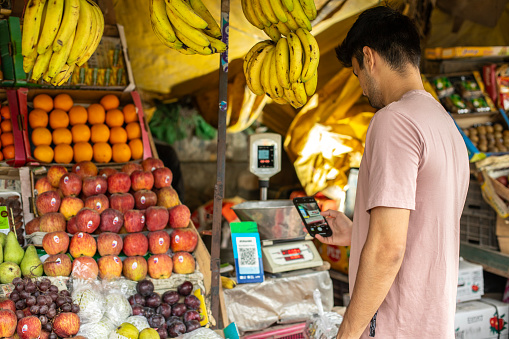 Side view of young man scanning QR code with smart phone and making online payment while buying fruit from market stall