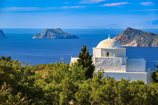 White Greek Orthodox chapel or church on hilltop of seashore on clear blue sky on sunny day. Clear sky, blue sea, distant islets in small harbour, rich greenery, green trees. Traditional religious building. Typical Greek landscape. Milos Greece