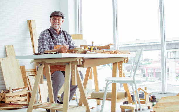 istock Senior old aging beard kind Caucasian man or carpenter wearing check shirt, apron, making DIY wooden toys for kids, smiling with happiness, sitting alone at indoor home. Retirement, Hobby Concept. 1690982793