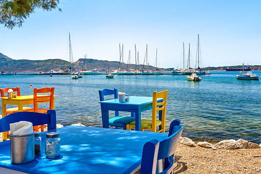 Colorful chairs and tables of Greek tavern by waterfront at sunny day. Vacations in Greece, summer, restaurant, outdoor dining, port, marina, yachts and boats, Mediterranean relax, Cyclades and Aegean islands. Selective focus, blurred background.