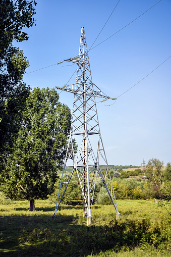 Metallic pylon of high-voltage power lines against blue sky. Located among trees with green foliage. The concept of power, transportation of electrical energy. Selective focus. Copy space.
