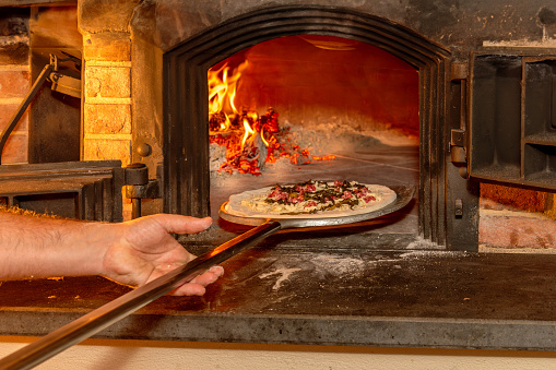 Hand of pizzailo puts pizza on a shovel in traditional wood-fired oven.