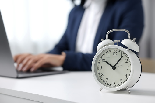 Stressful deadline. Alarm clock near woman working at white desk indoors, closeup. Space for text