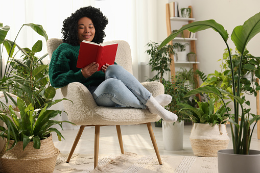 Relaxing atmosphere. Happy woman reading book on armchair surrounded by beautiful houseplants in room