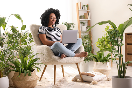 Relaxing atmosphere. Woman with laptop sitting on armchair surrounded by houseplants at home
