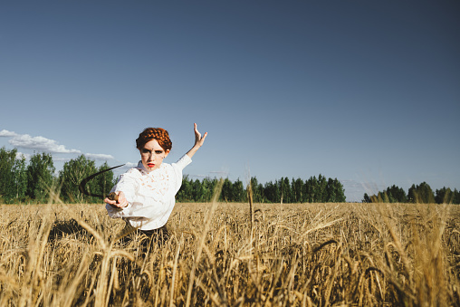 Slavic girl in a white dress and a black skirt with witch makeup stands in a field of rye and holds a terrible sickle in her hands. The concept of a mystical harvest.