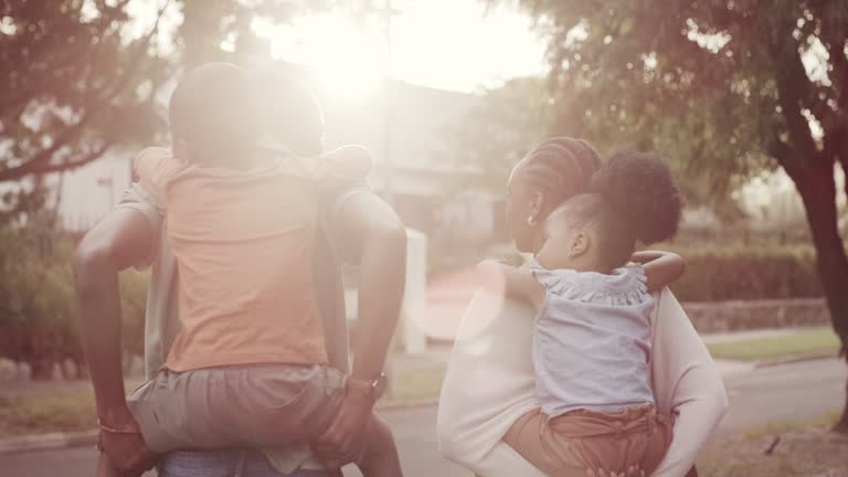 Black family, piggyback and walking together outdoor, bonding and having fun on sunset lens flare. Mother, father and carrying children on back with love, care and support to travel in neighborhood