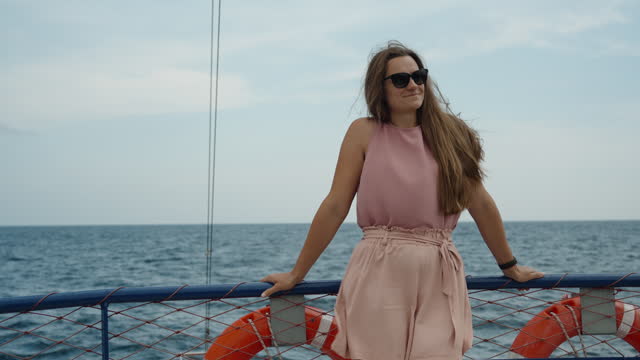 The young woman in pink shorts and a blouse stands on the upper deck of a cruise ship, smiling. slow motion