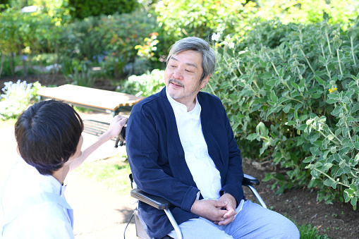 Young male caregiver smiling and talking with an Asian gray-haired elderly man in a wheelchair outdoors