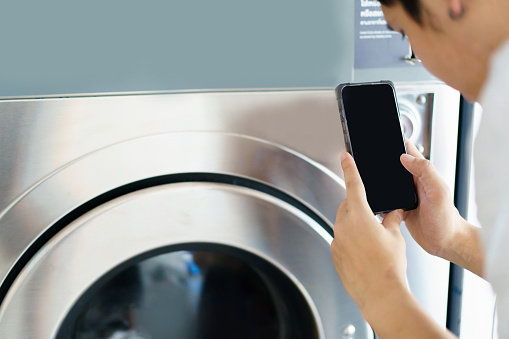 Isolated mockup white screen with clipping path. Asian man using self-service - automatic washing machine, Asian man using application on smartphone to access self service KIOS laundry machine.
