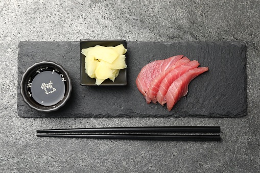 Tasty sashimi (pieces of fresh raw tuna) served with soy sauce and ginger slices on gray table, flat lay