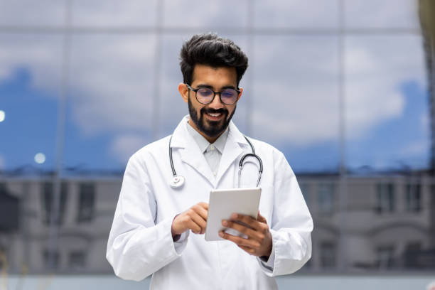 un jeune étudiant hindou à succès en blouse médicale blanche se promène à l’extérieur de la clinique, stagiaire médecin avec une tablette à la main, parcourant du matériel éducatif en ligne - trainee computer medical student student photos et images de collection