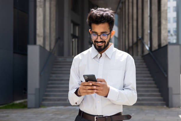 istock Closeup businessman walking city and using application on smartphone, a smiling Hispanic man holding a phone in his hands, browsing online pages, typing messages, and choosing in online store 1691078875