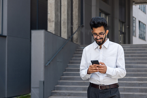 Young hispanic businessman walking outside office building in city, man in shirt holding phone, satisfied with achievement result freelancer, using online application.