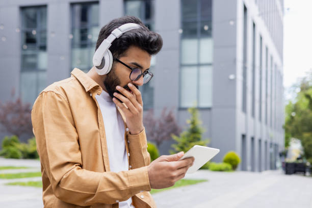 istock Young man with headphones and tablet computer sitting outside office building on park bench, Hindu man watching video online scary and sad using streaming app 1691085701