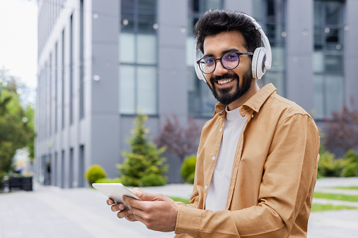 Portrait young Indian man with headphones and tablet watching online video sitting on bench near office building businessman smiling and looking at camera, portrait of satisfied online stream viewer