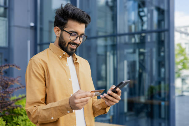 istock Young successful satisfied hispanic man smiling outside office building, businessman using app on phone for online shopping in online store, man holding bank credit card and smartphone 1691100134