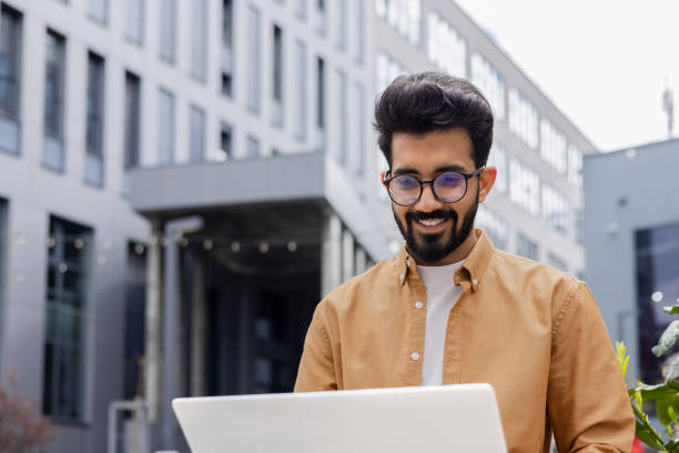 istock Young successful businessman working with laptop remotely sitting on bench outside office building, Hindu programmer in shirt smiling happy with achievement results and success 1691100146