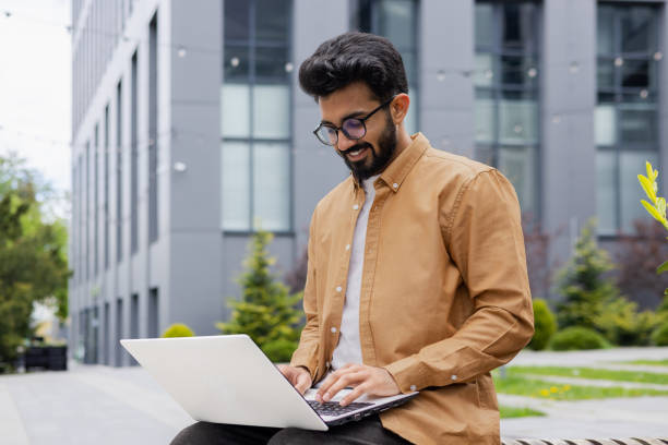 istock Young successful businessman working with laptop remotely sitting on bench outside office building, Hindu programmer in shirt smiling happy with achievement results and success 1691100149
