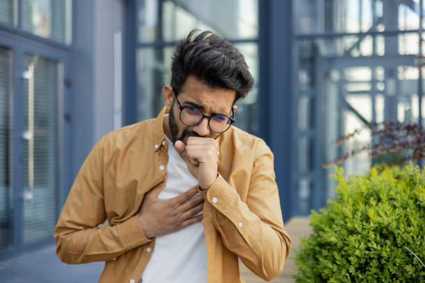 un enfermo se sienta en la calle, un hombre de negocios tose fuera de un edificio de oficinas, una mujer latinoamericana con ropa casual tiene un resfriado en un día soleado - coughing illness men latin american and hispanic ethnicity fotografías e imágenes de stock