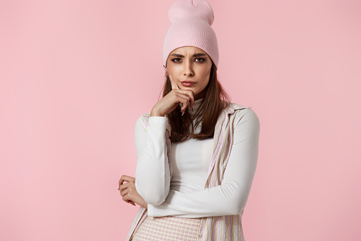 Thoughtful woman in pink hat keeps hand on chin looks pensively isolated on pastel pink background.
