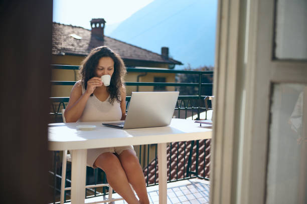 istock Beautiful woman drinking coffee while working remotely on laptop, sitting at table on the summer terrace of her house 1691155000