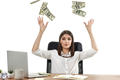 businesswoman working on laptop and throwing money while sitting on chair at desk. banknotes fly in the air