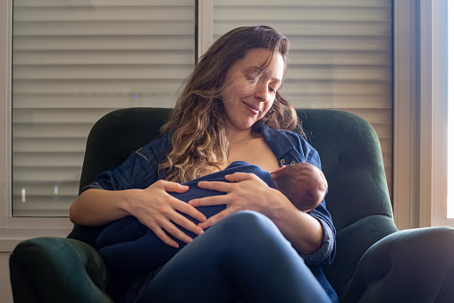 Brazilian mother breastfeeding her baby son while sitting on armchair at home.