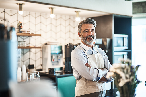 Portrait of a handsome male barista standing behind counter in a cafe. Mature man standing at cafe counter with arms crossed looking at camera..