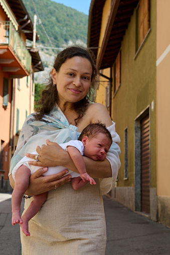 Beautiful happy woman holding her newborn baby, smiling looking at camera, standing in cobblestone alley in Italian medieval village - Canzo, Lombardy. Italy