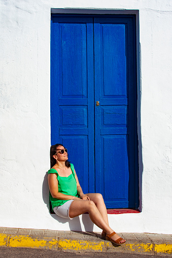 White woman with brown hair, wearing sunglasses, sitting on the step of a navy blue wooden door of a white house receiving the rays of the summer sun.
